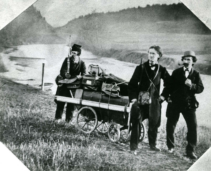 Gammelt foto av tre menn og en reisetralle i et landskap