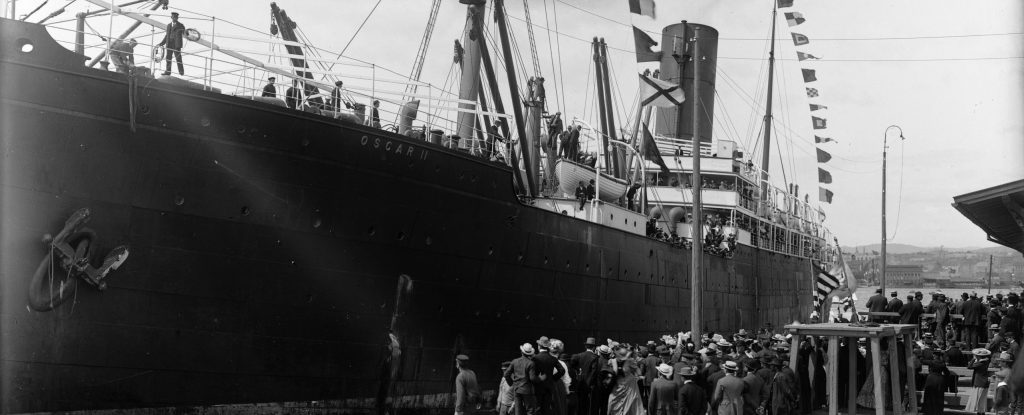 Svart-hvittfoto av stort skip ved en havn, med mange mennesker på havna og på skipet