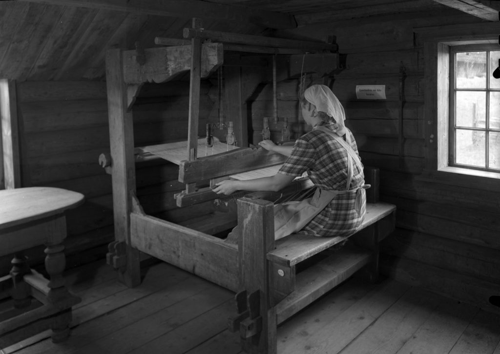 Foto av en kvinne ved en vevstol, innendørs i et eldre laftet bygg.