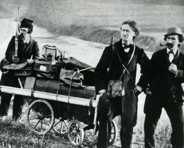 Gammelt foto av tre menn og en reisetralle i et landskap