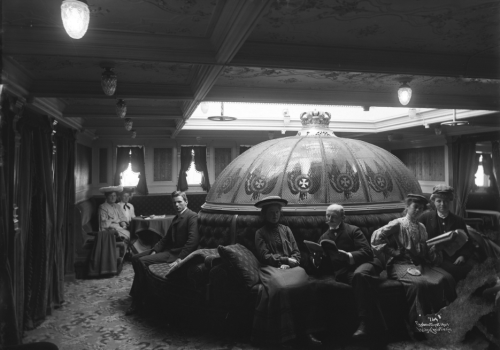 Utvandrere på vei til Amerika sitter i musikksalongen på første klasse på skipet S.S. Hellig Olav Svart-hvittfoto av pent kledde mennesker inne i en salong