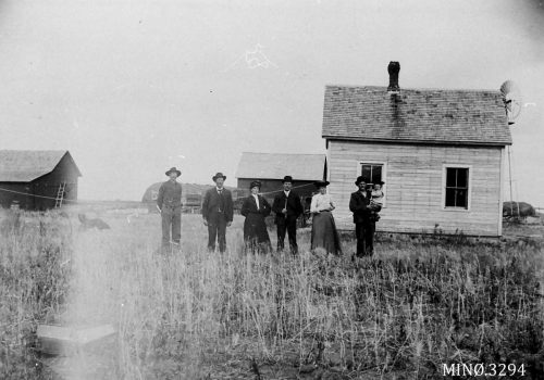 En Amerika-farm i USA ca. 1890-1935. Svart-hvittfoto av seks voksne og et barn foran et enkelt bolighus på en gård