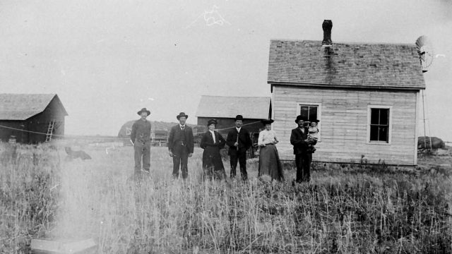 En Amerika-farm i USA ca. 1890-1935. Svart-hvittfoto av seks voksne og et barn foran et enkelt bolighus på en gård