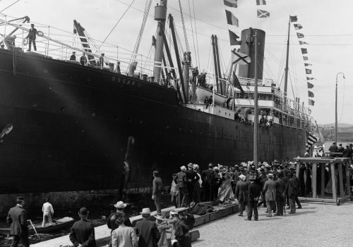 Emigrantskipet Oscar 2, fotografert av O. Væring en gang i tidsperioden 1890-1900. Svart-hvittfoto av stort skip ved en havn, med mange mennesker på havna og på skipet